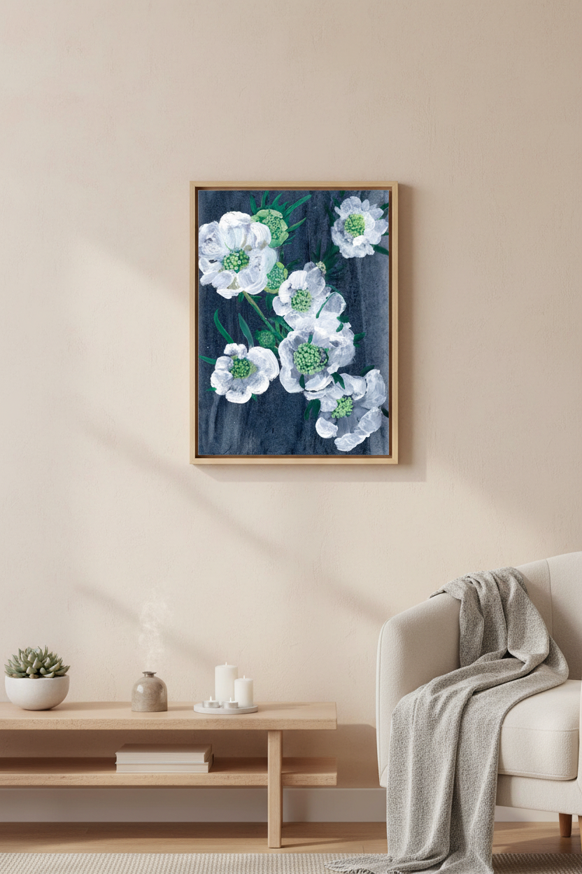 A framed floral wall art print featuring white flowers with green centers on a dark navy background, displayed in a modern living room setting above a light wood console table and next to a cream armchair with a gray throw blanket.