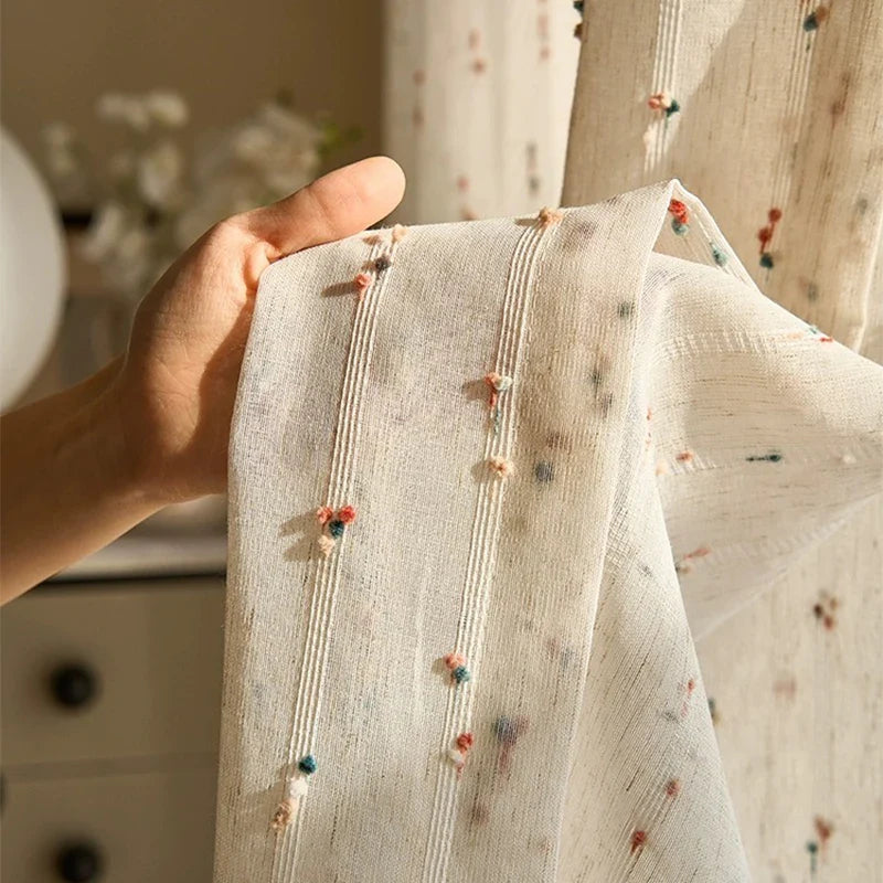 Hand holding a piece of light fabric with embroidered details in a softly blurred indoor setting.